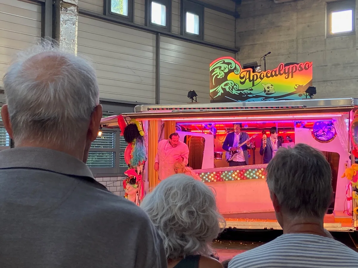 Ein bunter Jahrmarktstand, drinnen ein Gitarrist und zwei Schauspieler.